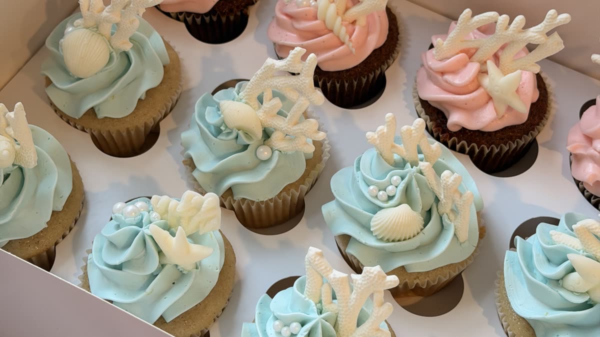 A box of beach-themed cupcakes with pink and blue frosting, seashells and coral toppers